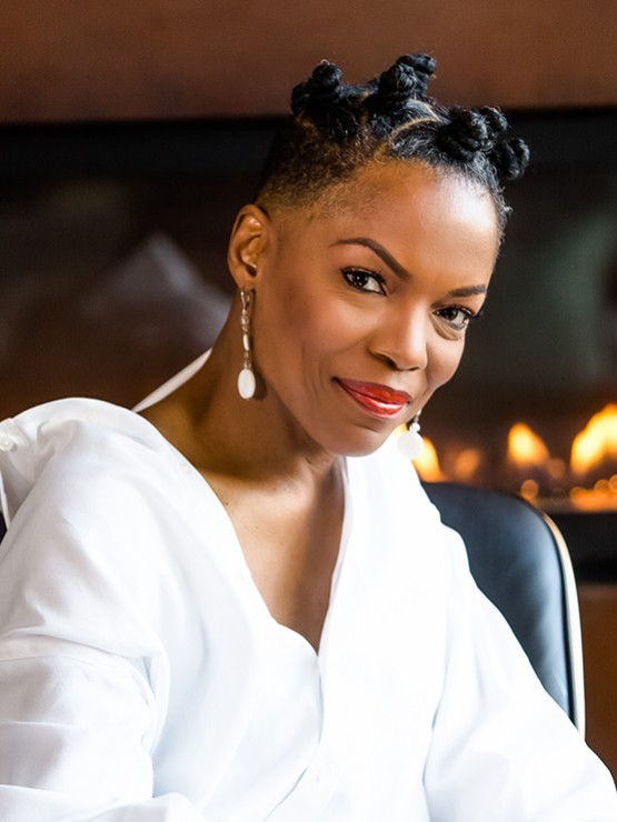 Nneena Freelon, a black woman in a white top, sits in front of a fire place and smiles.