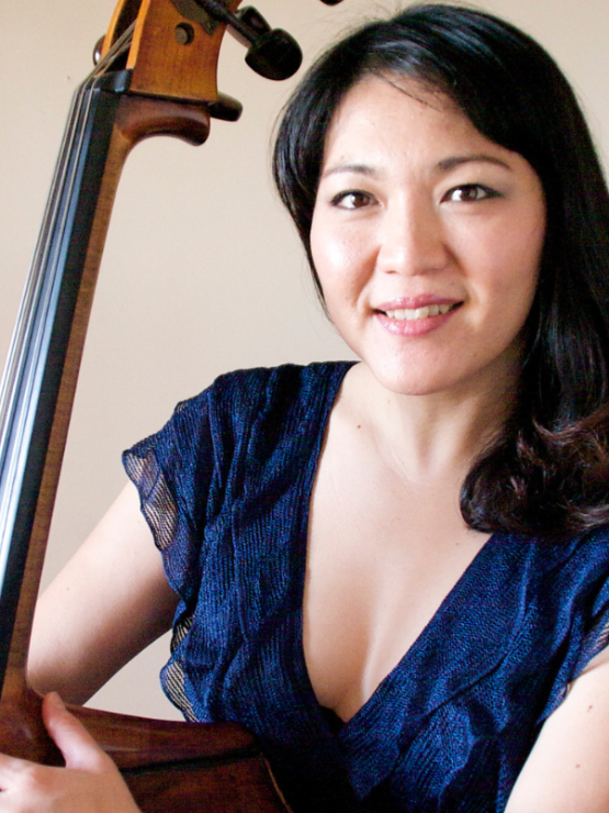 a long-haired japanese woman in a cap-sleeved dark blue dress. she stands with an arm around her double bass and smiles