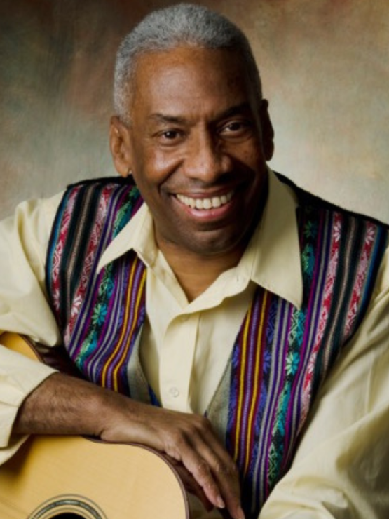 a middle aged black man with white hair and an ivory shirt with a patterned vest holds an acoustic guitar and smiles, slightly leaning forward