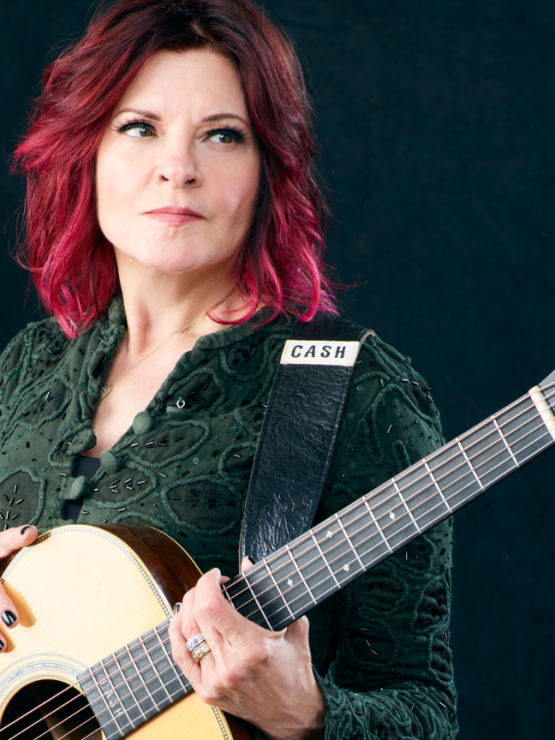 a white woman with vivid burgundy hair holds a guitar that reads CASH on the strap. She looks away out over the headstock of the guitar.