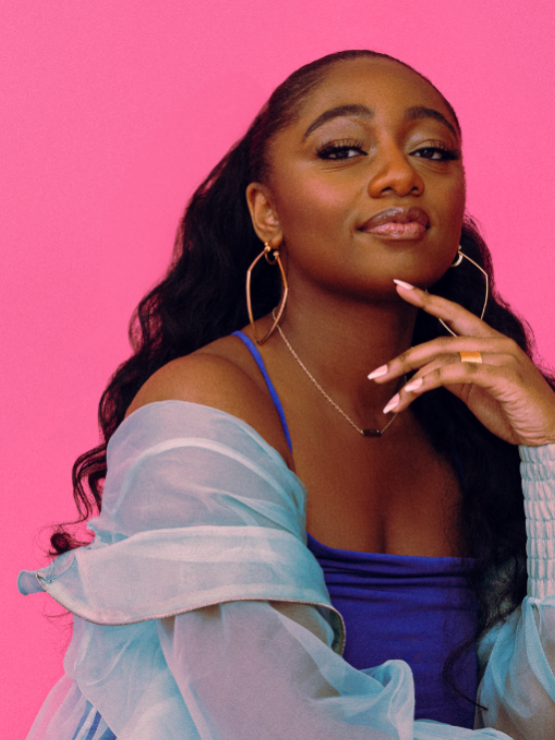 a black woman in her early 20s, with long hair and an ethereal wrap around her shoulders, raises a finger to her chin in front of a pink background