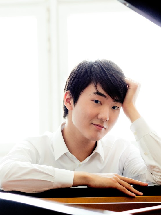 An Asian man with dark hair in a white shirt sits at a piano in front of a whitewashed window. 