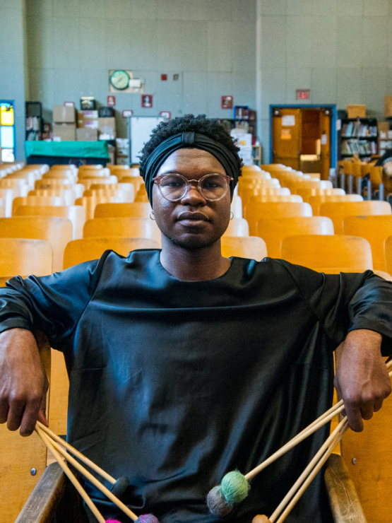 a nonbinary black person holds two sets of percussion mallets in their hands. they sit in a public school auditorium, with their elbows extended over wood-backed seats