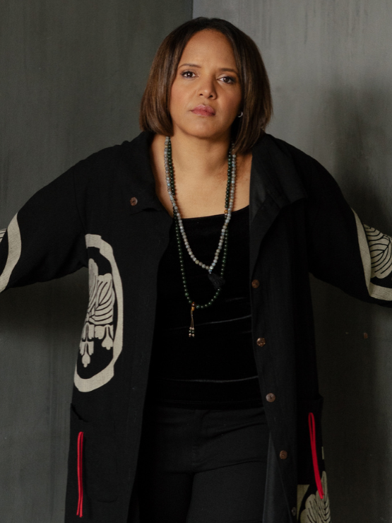 a Black woman in a black outfit topped by a long draped black jacket with abstract ivory medallion designs stands in a corner, with her hands on the concrete walls to either side of her.