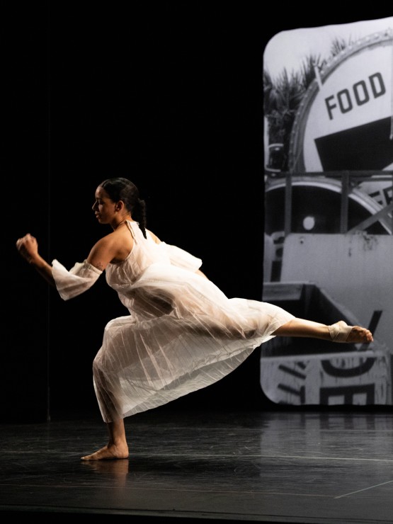 in front of a black and white photograph of roadway signs, two female dancers in sheer flowing ivory dresses move across the stage, one leaning forward on a bent leg, and the other with an arched back