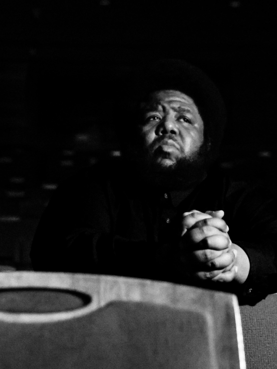 a low-contrast image of a black man in theater seating, his hands clasped and wrists resting on the seatback in front of him