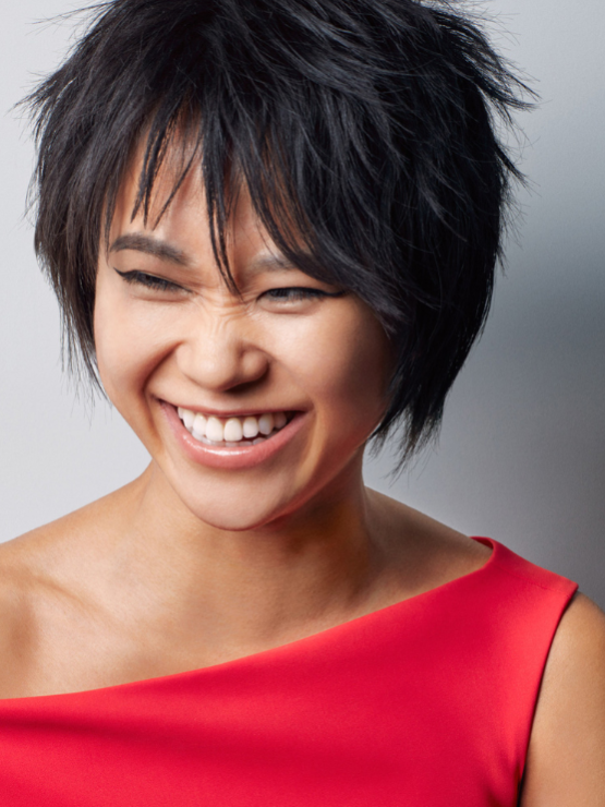 a glamorous chinese woman in her early 30s wrinkles her nose as she laughs. Her hair is in a chopped shag style, and she wears a red one-shoulder dress