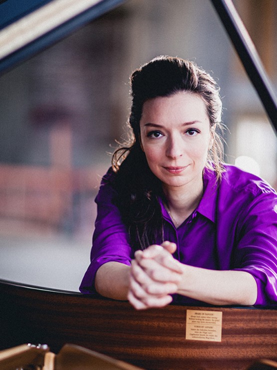 a dark-haired russian woman in her early 30s wearing a rich purple shirt leans over the edge of an open grand piano