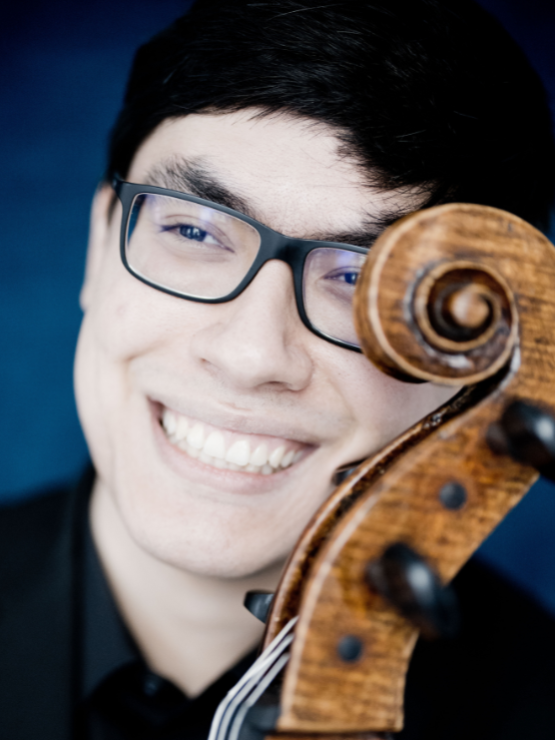 a man in his 20s, of eastern european and chinese heritage, smiles with the scroll of his cello curving across one cheek, partly covering his eye and eyeglasses
