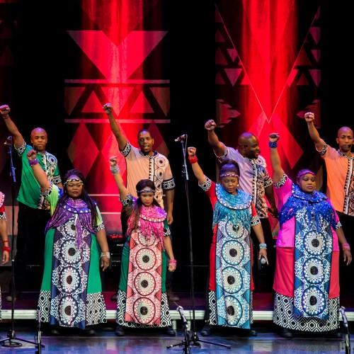 A group of singers in vibrant, colorful garments stand raising their right fists to the air in front of a red and black patterned background. 