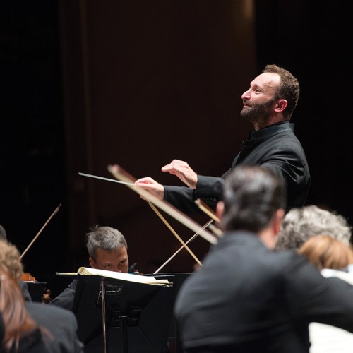 a conductor, focusing intently, pauses. he extends his baton flat and joins the thumb and forefinger of his other hand.