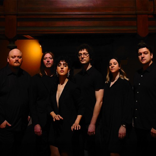 seven musicians in a performance space with warm amber light. some are turned to the side but all look at the camera
