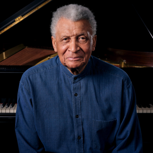 a multiracial man in his late 80s with medium brown skin and short gray and white hair stands with his back to the keyboard of a grand piano