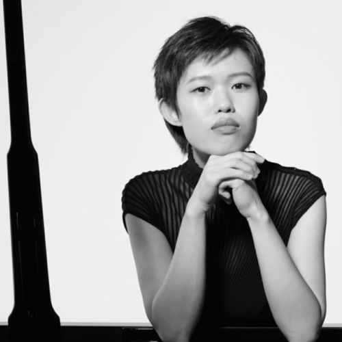 a chinese american woman in her 20s, with short hair in a pixie cut. she rests her elbows on a piano, her hands clasped in front of her face