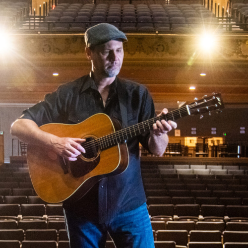 a white man in a cap plays an acoustic guitar on stage, an empty auditorium at his back