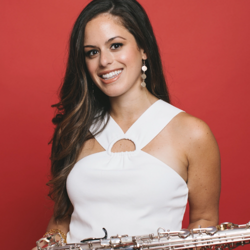 a smiling dark-haired woman in a white dress stands before a red background. She holds a saxophone in front of herself.