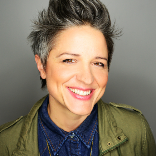 a woman with short salt-and-pepper hair swept up in a vertical style. she wears a denim shirt and an olive green jacket and smiles brilliantly