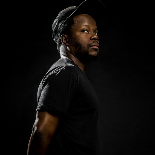 Ambrose Akinmusire, a black man in a black shirt and hat in front of a black background. 