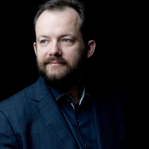 a bearded white man in his forties in a dark shirt and dark blue jacket, in a three-quarter view