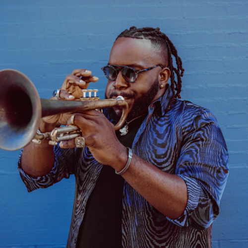 in the foreground the bell of a trumpet. the trumpeter, Marquis Hill, is a black man with hair in twists on the top and a beard. He wears sunglasses and a blue shirt and stands in front of a blue wall