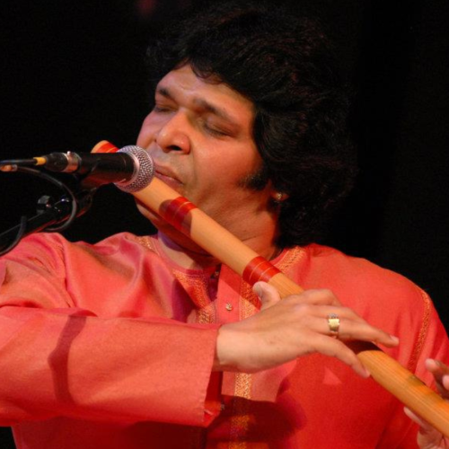 an indian man in an orange-red shirt plays a bansuri, a side-blown bamboo flute