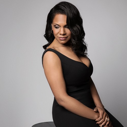 a black woman sits on a stool, looking over her shoulder with her hands in her lap. she wears a black gown.