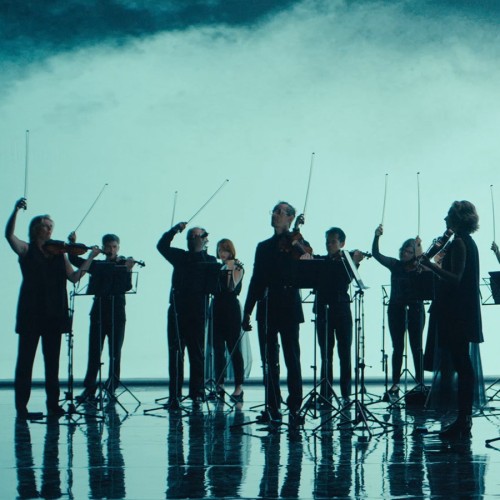 silhouetted before an aqua backdrop, a standing chamber orchestra raises their bows with a flourish