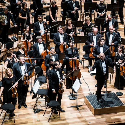 a symphony orchestra, in black tie concert dress, stand for a bow on a stage with a blond wood floor