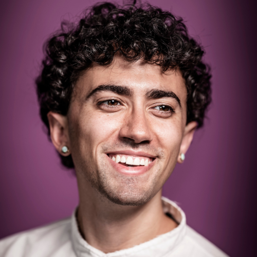 a white person in their late 20s with short brown curly hair, an ivory crewneck sweater, and stud earrings smiles in front of a purple background
