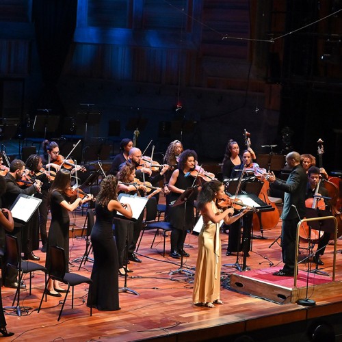 an orchestra with ethnically diverse members, standing, perform. A violin soloist, a woman in a long yellow dress, plays at the front.