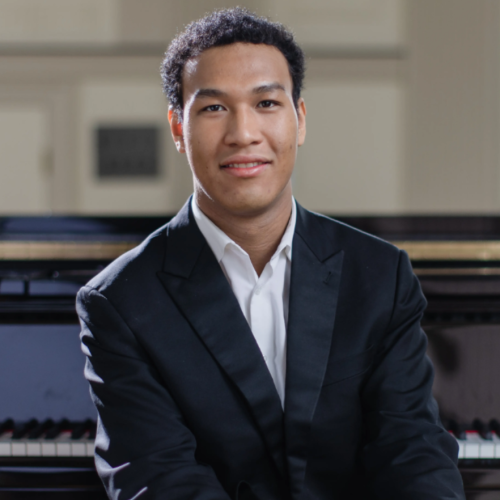 a young man in his early 20s with short dark curly hair and light brown skin sits in front of a grand piano in a brightly lit music room