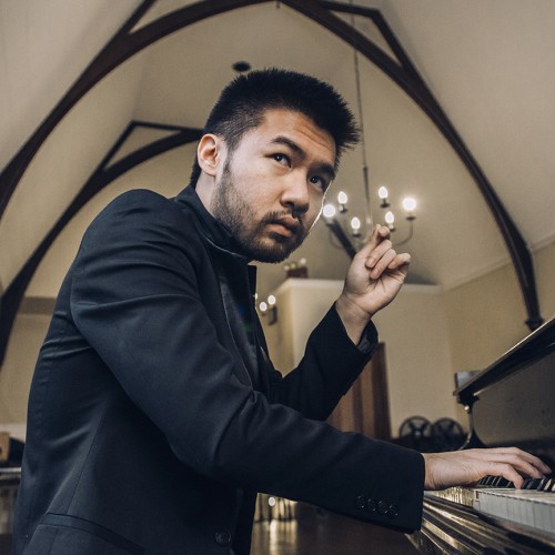 a young chinese-american man in a dark jacket plays the piano in a room with a vaulted ceiling. he has a scruffy short beard and one hand is lifted thoughtfully.