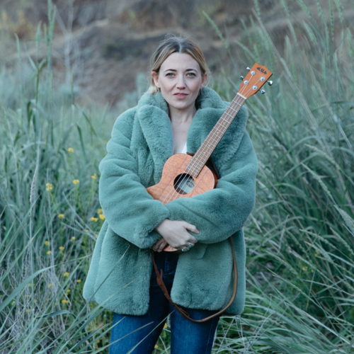 a blonde woman in a fuzzy green coat holds an ukulele in a field amid tufts of grass