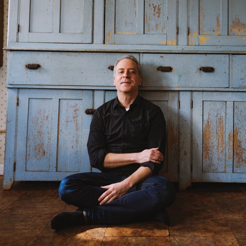 A white man in dark jeans and a black button-down shirt with the sleeves rolled up leans back against a rustic cupboard with flaking blue paint. 