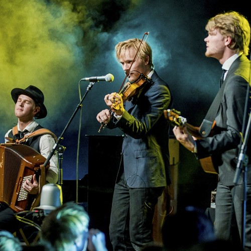Three men perform on stage with string instruments and microphones. Yellow smoke billows behind them. 