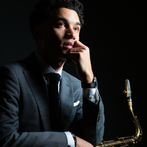 a puerto rican man with black hair wears a gray suit and faces right toward a light source. He holds a saxophone