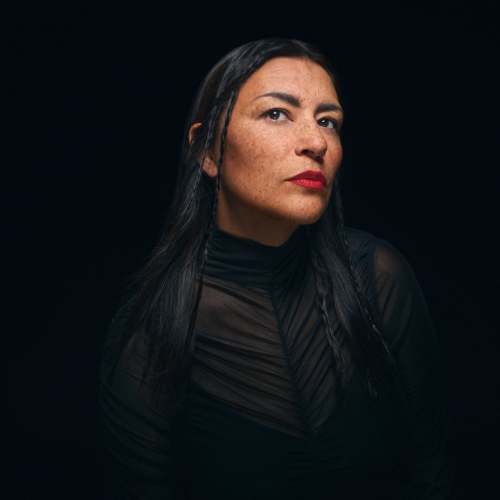 an indigenous woman in her 40s in black, against a dark background, in a three-quarter view. Her freckled face is framed by small braids, and she wears vivid red lipstick