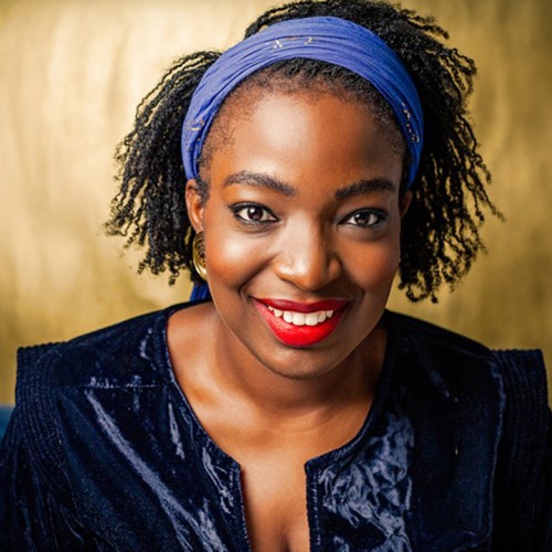 a warmly smiling black woman with a wide light blue headband. The lighting is golden, and she wears a chic navy jacket with a bright red lipstick