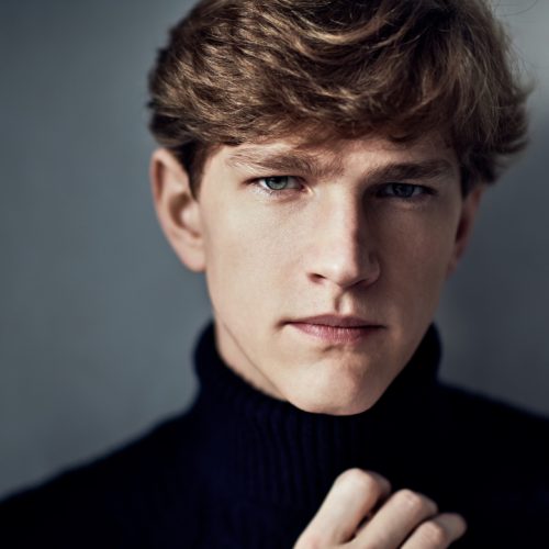 a white man in his 20s with sandy brown hair. he wears a black turtleneck. one hand rests on his chest, with fingers curled