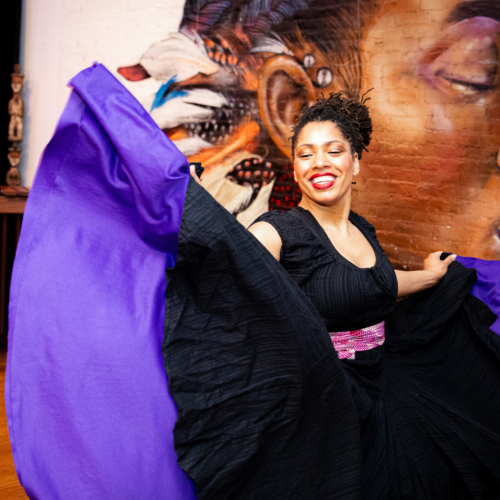 a multiracial black woman in a black dress swirls her skirt, revealing a wide purple hem. she stands in front of a mural depicting a brown-skinned elder whose face we cannot see