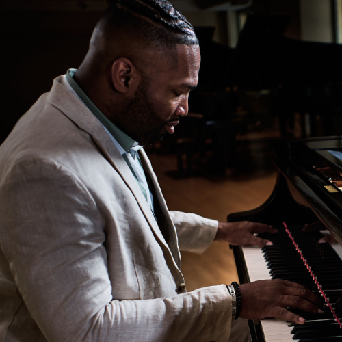 a black man in a light gray suit looks down at his hands as he sits sideways at a black piano