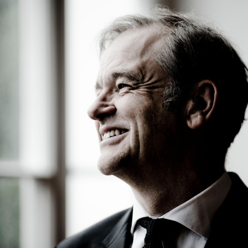 a white english man in his sixties turns to the side, smiling. the lighting is bright but cool, and he wears a jacket and tie