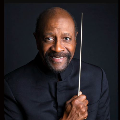 a black man in his 60s, with a goatee and a bald head, wears a black turtleneck against a black background. he holds a white conductor's baton vertically, close to the side of his face