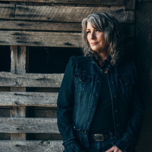 a white woman with wavy gray hair stands in front of a rustic wall. She wears a dark ruffled denim jacket and dark jeans with a scrolled belt buckle.