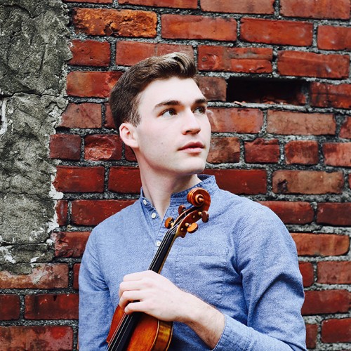 a young white man holds a violin and looks off and to his left. he stands outdoors in front of a brick wall.