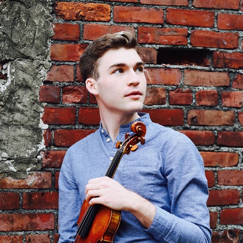 Alexi Kenney a young brown haired white man in a blue shirt stands in front of a brick wall holding a violin. 