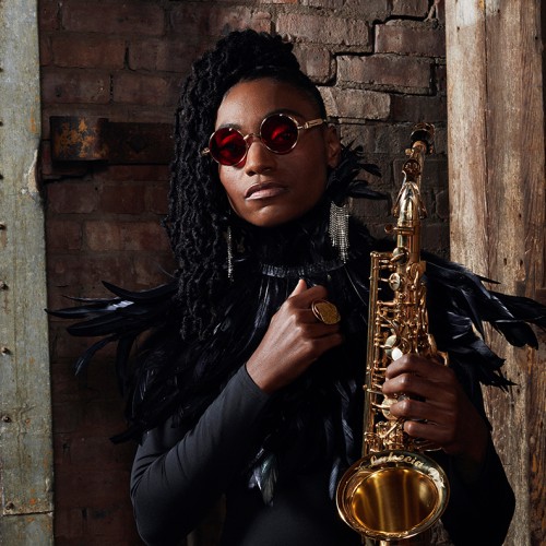 In front of a brick wall, a black woman holds an alto saxophone. She wears heavy gold-framed round sunglasses and black-feathered epaulets on her shoulders. 