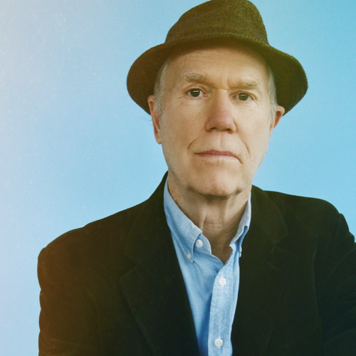 an older white man wearing a blue shirt, a dark jacket, and a hat stands before a light blue background