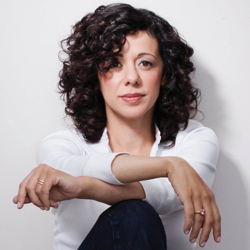 a curly-haired woman in a white shirt sits with her arms casually crossed