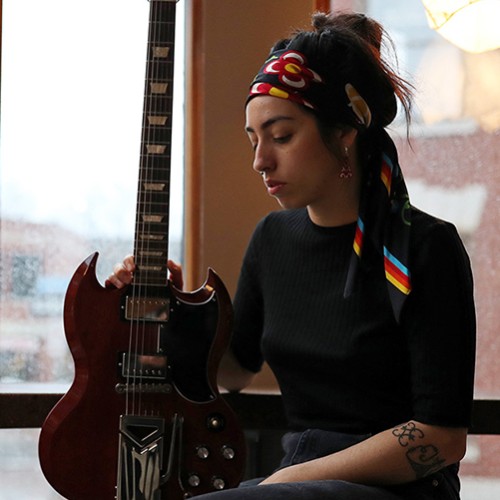 an Indigenous person in her 20s wearing a headwrap with stripes and flowers sits in a windowed corner, looking down at the bass guitar she holds
